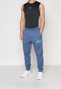 Zwarte mouwloze top en blauwe trainingsbroek met een wit Nike-logo. De broek heeft een elastische tailleband en boorden, gemaakt van een zachte stof.