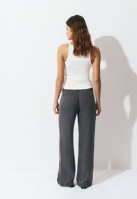 Woman with wavy brown hair stands facing a white wall wearing a white tank top and loose-fitting dark gray pants.