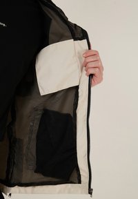 Lichte jas met zwarte meshvoering en een crèmekleurige zak. De stof is glad, met een ritsluiting en contrasterende texturen.
