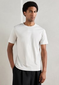 Svetlo šedé tričko s krátkym rukávom vyrobené z jemnej bavlny. Má okrúhly výstrih a subtílne logo na hrudi. Kombinované s čiernymi nohavicami.