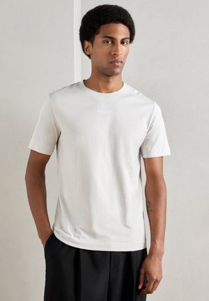 Helles graues kurzärmliges T-Shirt aus weichem Baumwollstoff. Mit Rundhalsausschnitt und dezentem Logo auf der Brust. Getragen mit schwarzen Hosen.