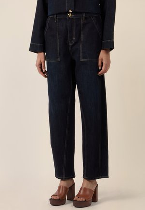 Woman wearing dark blue wide-leg jeans with visible stitching and brown open-toe platform heels against beige background.