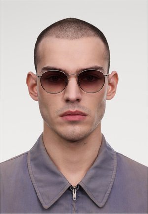 Lunettes de soleil rondes avec des verres teintés foncés et des montures en métal argenté, portées par un modèle dans une veste grise sans col avec une fermeture éclair.