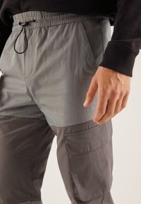 Lekki, dwukolorowy materiał spodni w odcieniu szarym z elastycznym pasem, bocznymi kieszeniami oraz teksturowaną tkaniną. Posiada widoczne szwy oraz kieszeń cargo.