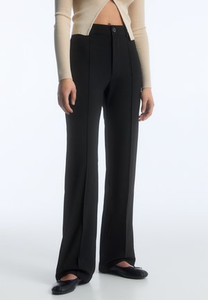 Personne portant un pantalon noir taille haute à jambes larges avec coutures frontales, associé à des ballerines noires et un pull court côtelé beige.