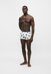 Мускулест мъж стои бос, облечен в бели боксерки с лента на талията Polo Ralph Lauren в синьо и щампи на мечки на бял фон.