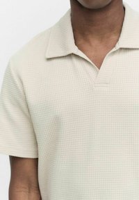 Lyhyt hiha polo-paita vaaleanbeigessä värissä, jossa on viistopintaista, vohvelikangasta. Sisältää kauluskannan ja osittaisen nappilistan.