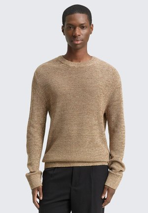 Giovane uomo che indossa un maglione a maglia testurata beige e pantaloni neri, in piedi contro uno sfondo grigio chiaro uniforme.