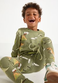 Ragazzo sorridente con capelli ricci, vestito con pigiama a stampa di dinosauri verde e sneakers, seduto sul pavimento contro uno sfondo bianco.