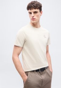 Νεαρός άνδρας στέκεται με τα χέρια στις τσέπες, φορώντας μπεζ κοντομάνικη μπλούζα και ανοιχτό καφέ παντελόνι με μαύρη ζώνη.