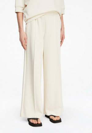 Osoba nosząca luźne kremowe spodnie z szerokimi nogawkami i czarne sandały na palce z kwadratowym noskiem, stojąca na białym tle.