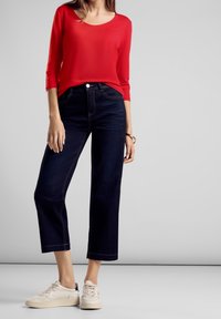 Rotes Langarmshirt kombiniert mit dunkelblauen, knöchellangen Jeans. Weiße Sneakers mit beigen Akzenten. Einfaches Design, glatte Textur des Stoffes.