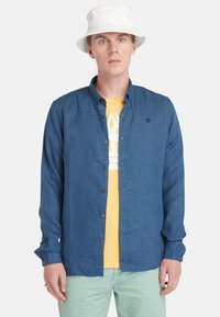 Camisa azul de botones en una tela ligera, con mangas largas enrolladas en los puños, superpuesta sobre una camiseta gráfica amarilla, combinada con pantalones verde menta.