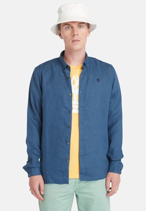 Camicia blu a maniche lunghe in tessuto leggero, maniche arrotolate ai polsi, indossata sopra una t-shirt gialla con grafica, abbinata a pantaloni verde menta.
