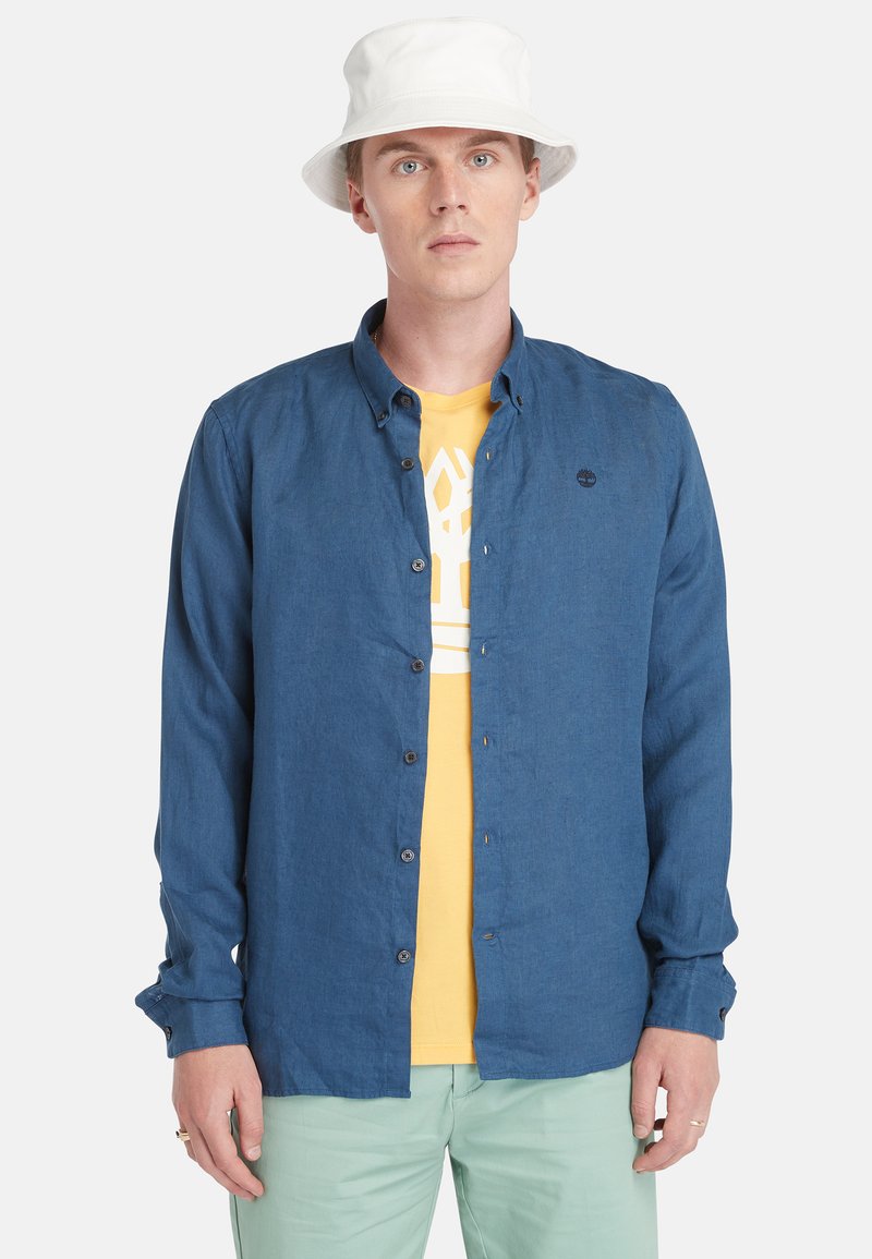 Camisa azul de botones en una tela ligera, con mangas largas enrolladas en los puños, superpuesta sobre una camiseta gráfica amarilla, combinada con pantalones verde menta.