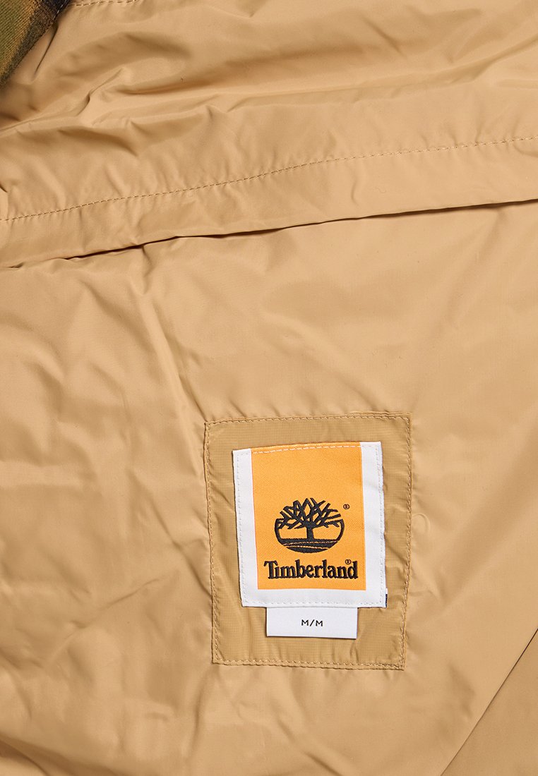 Tessuto beige con patch del logo Timberland cucito in arancione e bianco, con un emblema di albero nero e l'etichetta di taglia M/M sottostante.