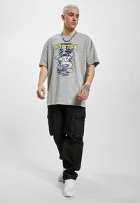 Grey oversized t-shirt with a space-themed graphic and “NO RETURN” text. Black cargo pants with side pockets. White sneakers. Silver chain necklace.