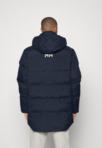 Marineblå pufferjakke med hætte, vandret quiltet design og et logo på ryggen. Nederste kant er længere bagpå.