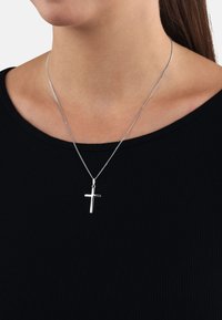 Silbernes Kreuz auf einer dünnen Kette, das am Hals ruht. Der Hintergrund zeigt ein schwarzes geripptes Oberteil. Glatte Textur mit polierter Oberfläche.