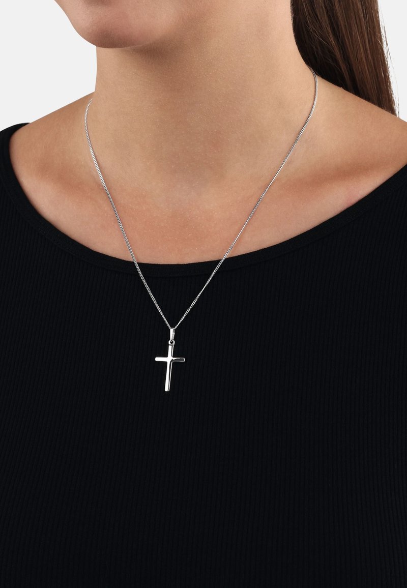 Silbernes Kreuz auf einer dünnen Kette, das am Hals ruht. Der Hintergrund zeigt ein schwarzes geripptes Oberteil. Glatte Textur mit polierter Oberfläche.