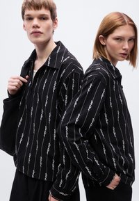 Μαύρα ζακέτες με φερμουάρ, με κάθετες λευκές γραμμές και μοτίβα με κείμενο. Μαλακό ύφασμα, λεπτομέρεια γιακά, φορεμένα από δύο μοντέλα μπροστά σε απλό φόντο.