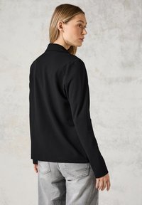 Blazer nero a maniche lunghe con colletto, texture liscia, cuciture dettagliate e vestibilità leggermente aderente, abbinato a pantaloni grigi chiari.