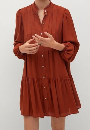 Femme portant une robe à manches longues couleur rouille avec boutons, plis et col à volants, les mains positionnées près de la poitrine.