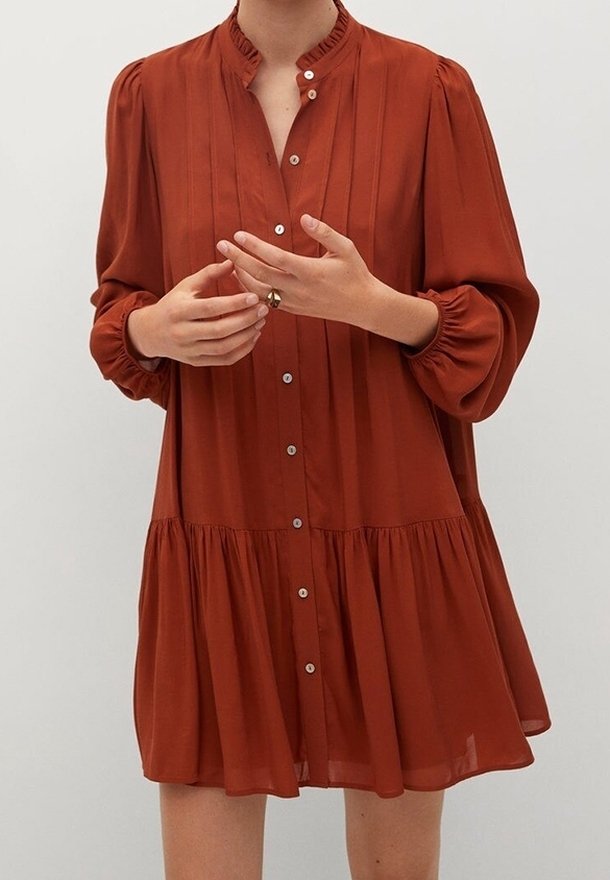 Femme portant une robe à manches longues couleur rouille avec boutons, plis et col à volants, les mains positionnées près de la poitrine.