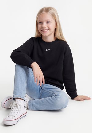 Ung pige med langt blondt hår iført en sort Nike sweatshirt, lyseblå jeans og hvide sneakers, siddende på gulvet og smilende.