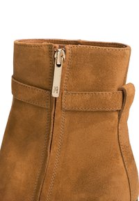 Gros plan d'une botte en daim marron avec une fermeture éclair verticale et un détail de sangle cousue près du bord supérieur.