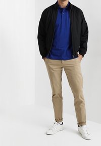 Veste bomber noire, polo bleu, chinos beiges, sneakers blancs. Tissu texturé, col standard, et poignets élastiques visibles. Design simple.