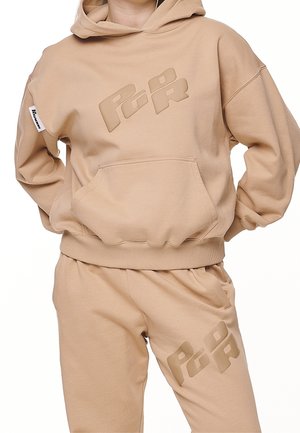 Beżowa bluza z kapturem i dopasowane spodnie dresowe z logo "PQR" na piersi i udzie, noszone przez osobę z rękami na biodrach.