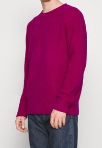 Maglione fucsia lavorato a maglia con scollatura rotonda, polsini e orlo a coste, realizzato in un tessuto morbido e testurizzato. Abbinato a jeans scuri casual.