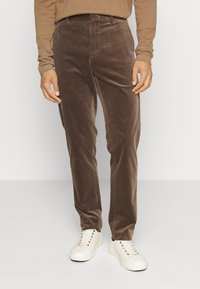 Pantalon en velours côtelé marron, coupe droite, avec un motif texturé. Porté avec des baskets blanches. Design simple et classique.