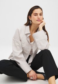 Camisa de botones a rayas en tela gris claro y blanca, con corte relajado y puños anchos, combinada con pantalones negros y zapatos negros sencillos.