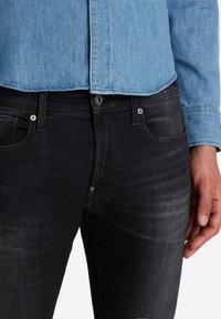 Jeans de mezclilla oscura con corte ajustado, que presentan un sutil efecto de desgaste y un estilo de cinco bolsillos. La tela tiene una textura suave con detalles de botones en contraste.