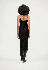 Sieviete ar krullainiem matiem, tērpta melnā midi kleitā ar aukliņām un melnām augstpapēžu ceļa auguma zābakiem, stāvot pret vienkāršu fonu.