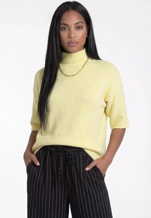 Kobieta o długich, prostych czarnych włosach, ubrana w żółty sweter z krótkim rękawem i golfem oraz czarne spodnie w cienkie paski, trzyma ręce w kieszeniach.