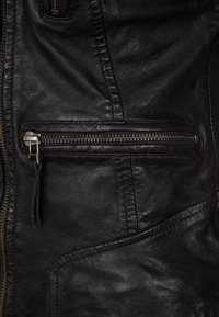Giacca di pelle nera con una tasca con zip diagonale, superficie testurizzata e dettagli di cucitura visibili. Accentata da hardware metallico.