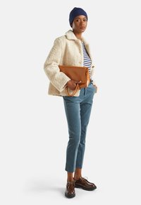 Mujer con una chaqueta de forro polar color crema, camiseta a rayas, gorro azul, pantalones azules y zapatos marrones, sosteniendo un gran bolso de mano de cuero marrón.