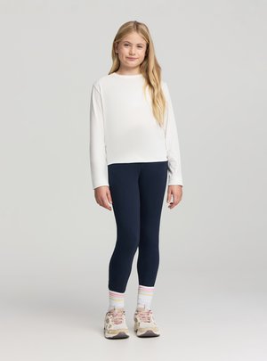 Ragazza in piedi che indossa una maglietta a maniche lunghe bianca, leggings blu navy, calzini bianchi con strisce rosa e scarpe da ginnastica beige su uno sfondo chiaro e uniforme.