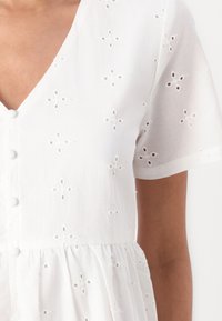 Bílé bavlněné šaty s výstřihem do V, krátkými rukávy, knoflíkovým zapínáním, květinovou výšivkou a elastickým pasem.