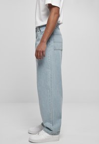 Persona che indossa jeans a gamba larga blu chiaro, sneakers bianche e una camicia corta bianca, in piedi di lato su uno sfondo semplice.