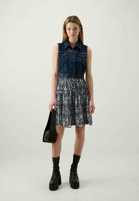 Tânără îmbrăcată cu o vestă din denim fără mâneci, fustă cu model, cizme negre cu platformă, ținând o geantă neagră în fața unui fundal simplu.