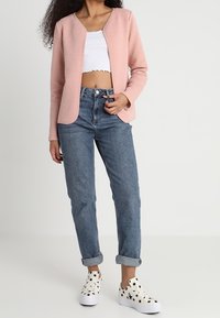 Světle růžová texturovaná bunda, bílý krátký top, džíny a bílé tenisky s černými tečkami. Bunda má otevřený přední díl a dlouhé rukávy.