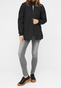 Kvinna iklädd en svart kviltad jacka, grå smala jeans och svarta sneakers med bruna sulor, stående med en hand på jackans dragkedja.