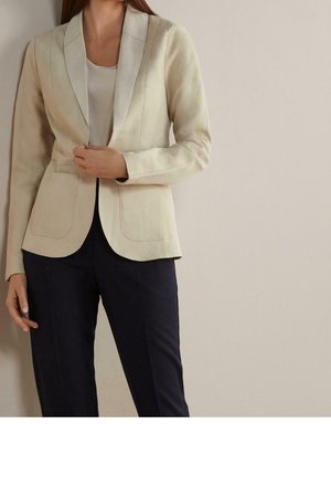 Donna con blazer beige sopra una maglietta bianca e pantaloni scuri, in piedi contro una parete chiara e semplice, che sistema il blazer con una mano.