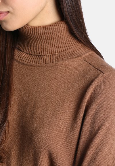 Femme portant un pull à col roulé côtelé marron avec de longs cheveux raides et foncés, montrée du cou à la partie supérieure de la poitrine et à l'épaule.