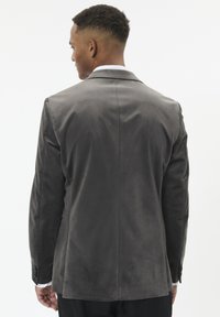 Mand stående med ryggen til iført en grå velourblazer, hvor hvide skjortermærker er synlige, og sorte bukser mod en simpel hvid baggrund.