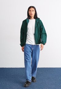 Grønn lett jakke over en hvit skjorte, kombinert med blå løse jeans og svarte sko. Jakken har en avslappet passform og elastiske ermer.